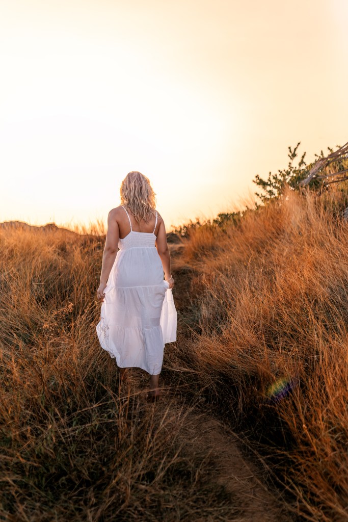 A woman wearing a white dress walking along a path in a field of tall grass during sunset.