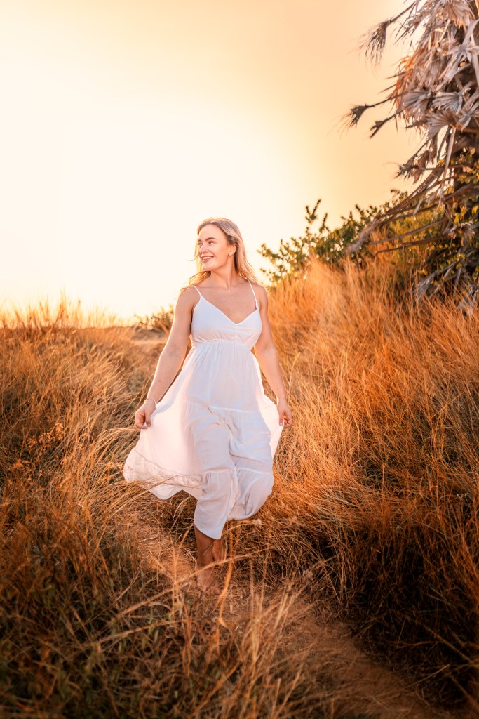 A smiling woman in a white dress walks through tall golden grass, with a warm, colorful sunset in the background.