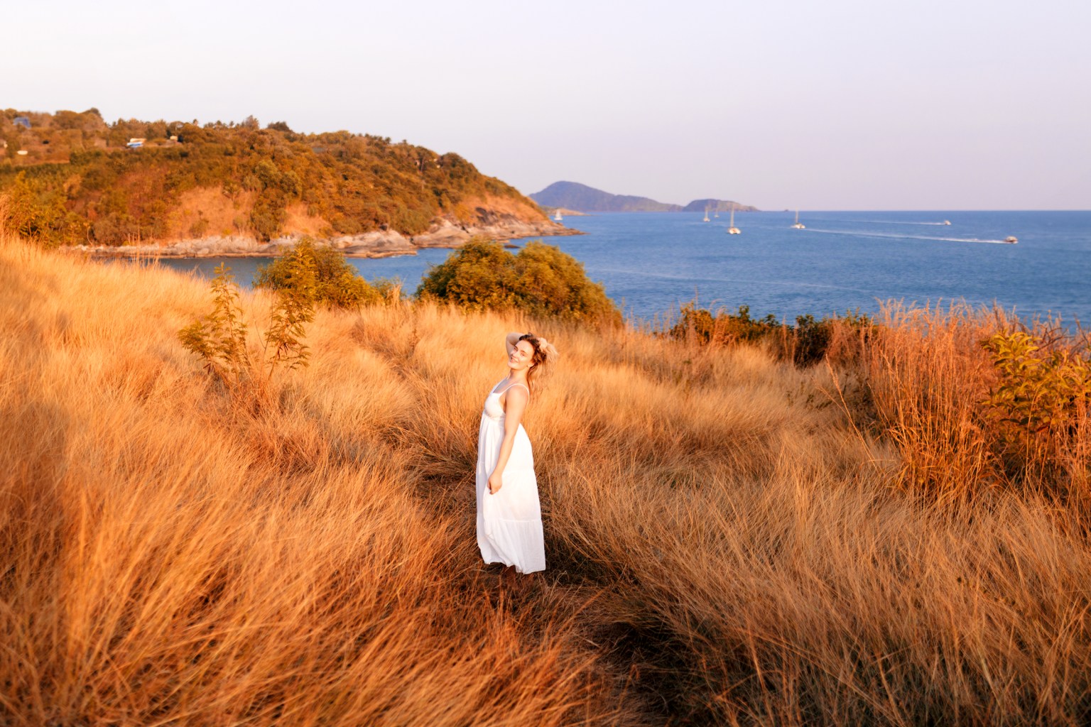 A woman in a white dress standing in tall golden grass with a scenic coastal view in the background, featuring blue water and distant hills.