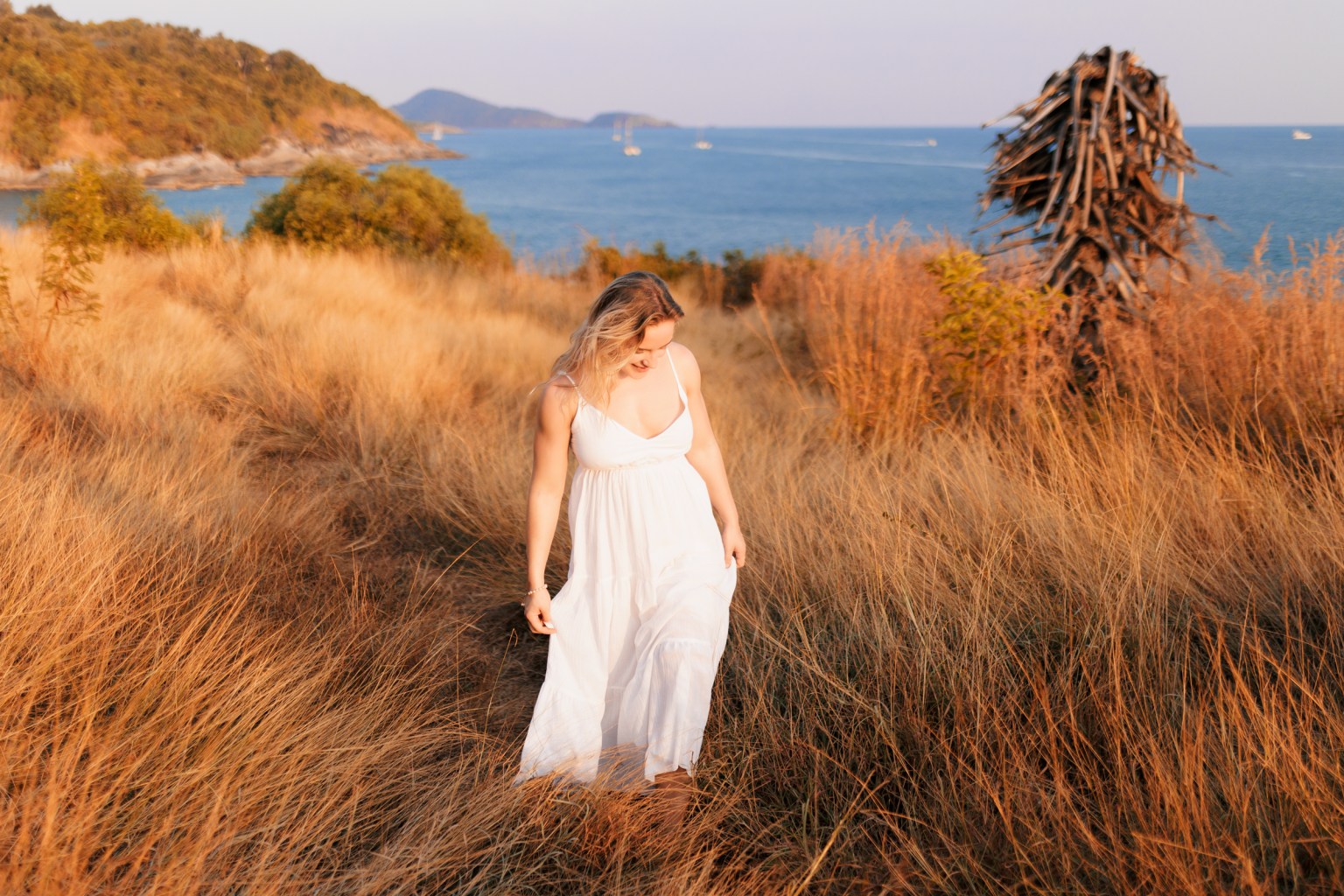A woman in a white dress walking through golden grass with a coastal background and a serene sea view.