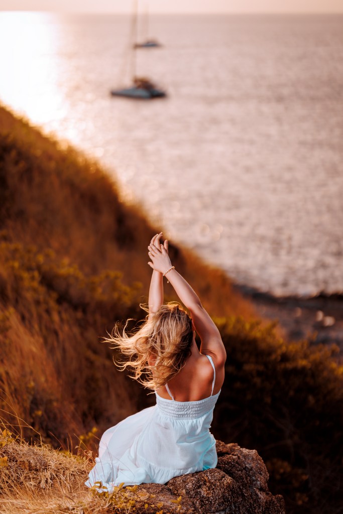 A woman in a light blue dress sits on a rock, enjoying the view of the ocean at sunset with sailboats in the background.