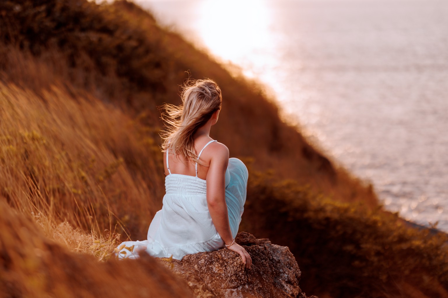 A woman sitting on a rock by the sea, looking out at the ocean during sunset, with grass and foliage in the background.