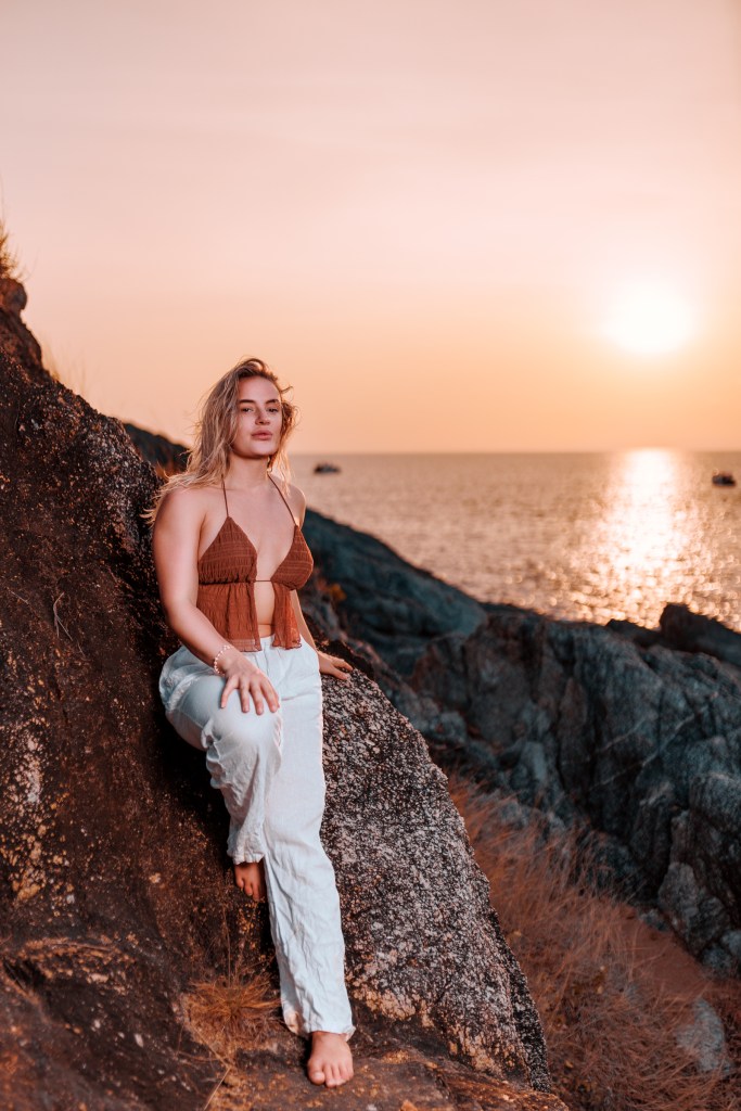 A person sitting on a rock by the seaside during sunset, wearing a brown top and white pants, with the sun setting over the ocean in the background.