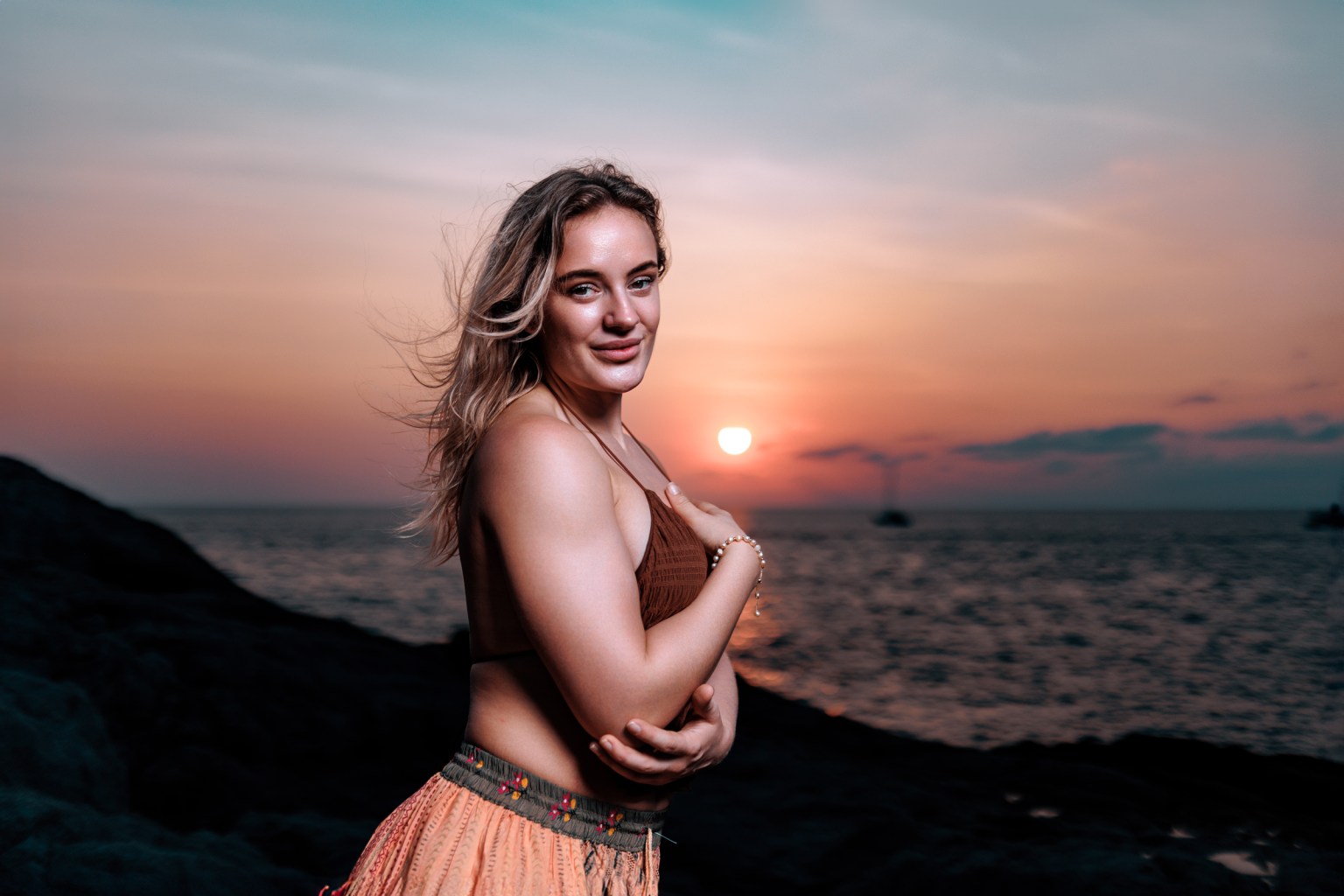 A woman standing on a rocky beach during sunset, with the sun setting over the ocean in the background. She is wearing a brown top and floral skirt, and has long, flowing hair.