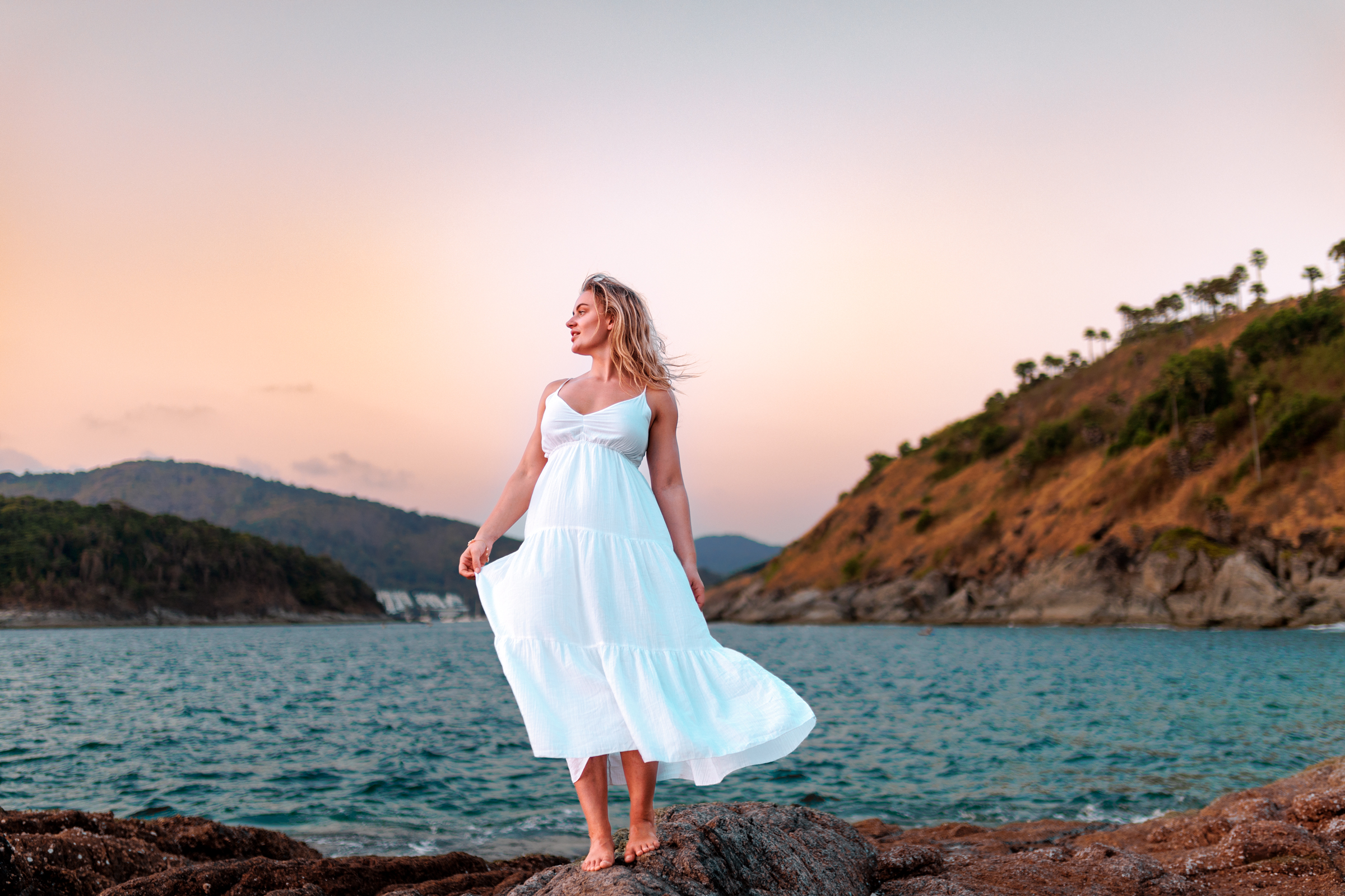 A woman in a flowing white dress stands on rocks by the sea at sunset, with hills in the background.
