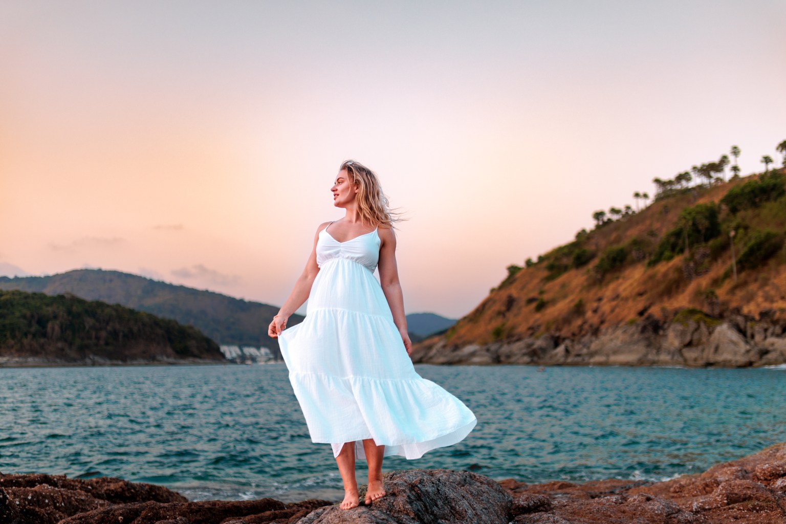 A woman in a white dress standing on rocks by the sea during sunset, with mountains in the background.