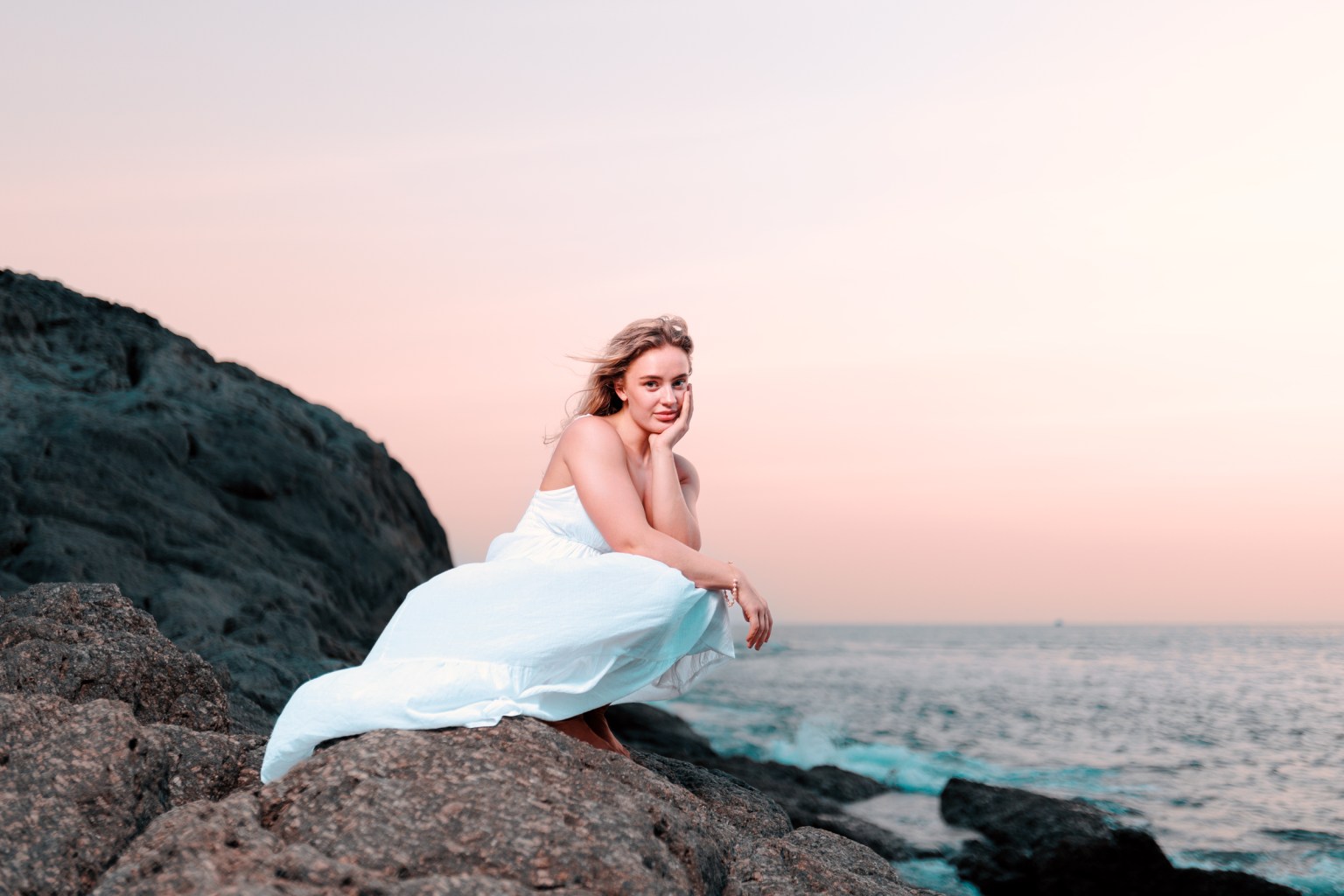 A woman in a white dress sitting on rocks by the sea during sunset, with a soft pink sky in the background.