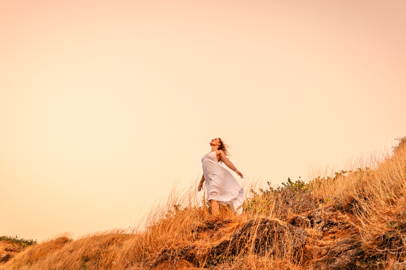 A woman in a flowing white dress standing on a hillside, with her arms outstretched, against a soft pink sky.