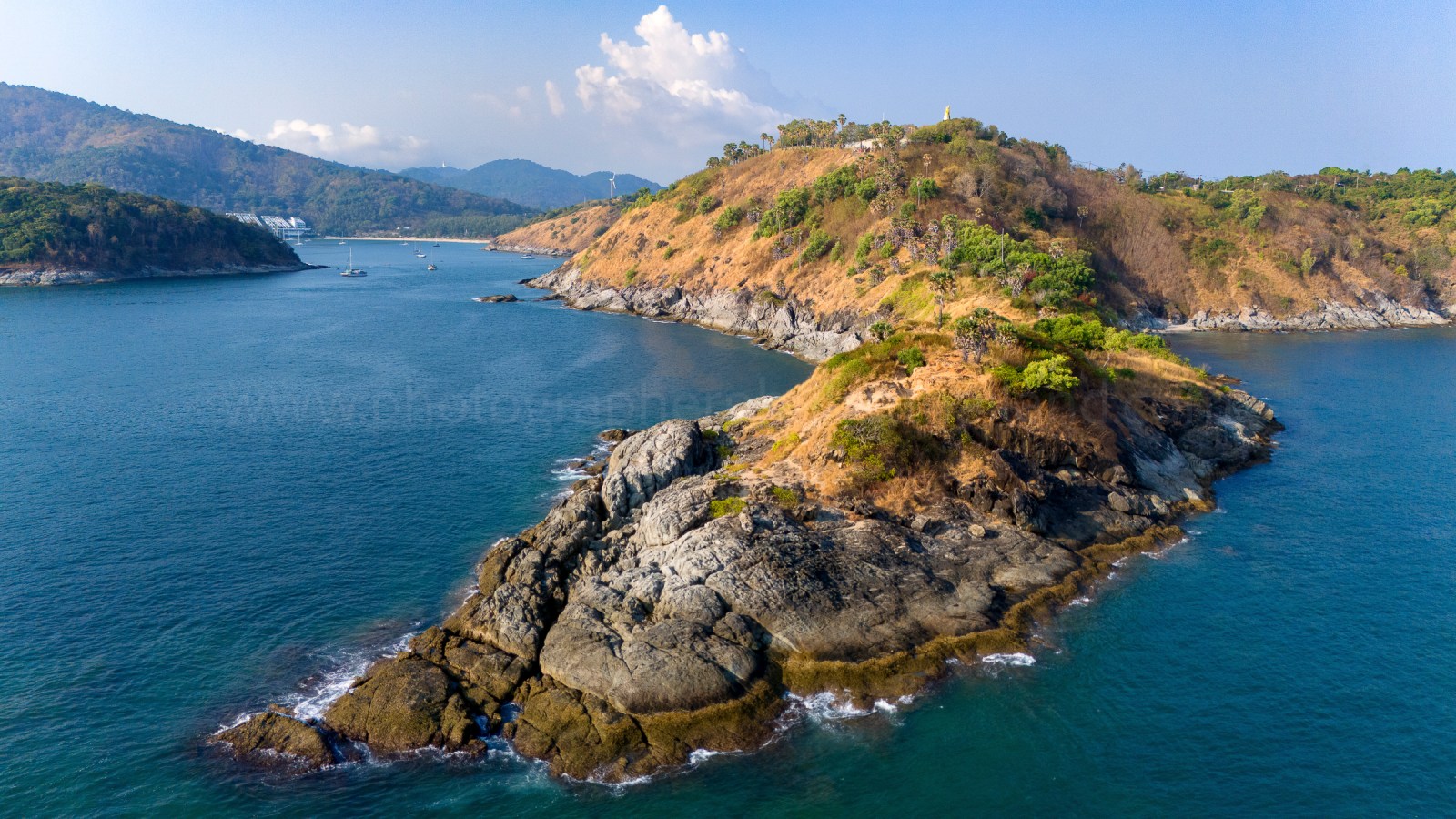 Aerial view of a rocky coastline with green hills and calm blue waters, featuring boats in the distance.