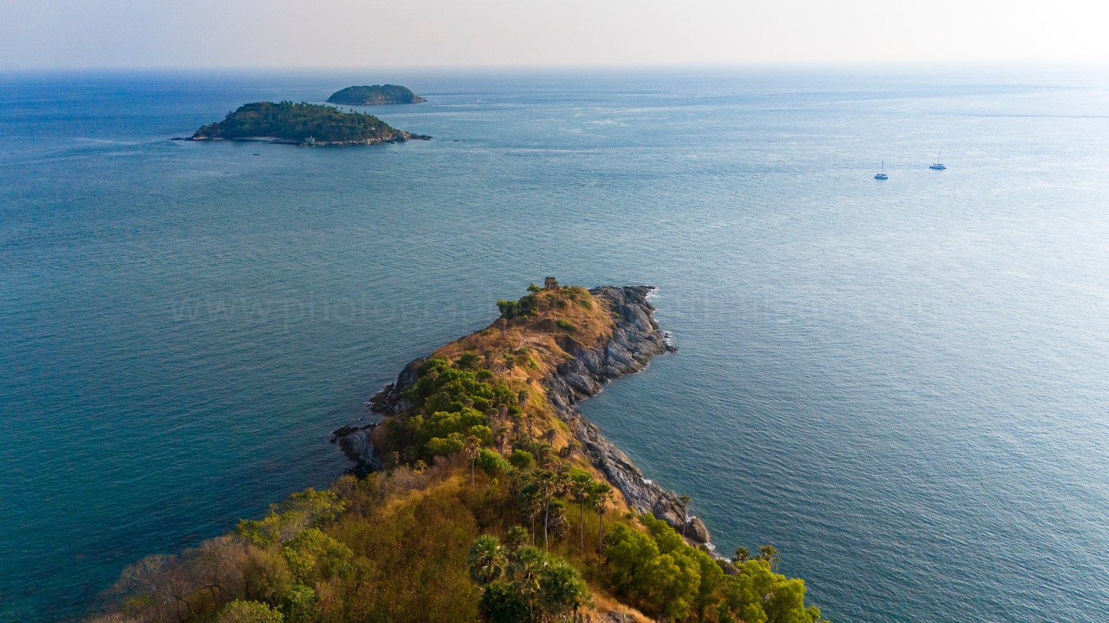 Aerial view of a rocky peninsula extending into the blue sea, with lush greenery on the shore and small islands in the distance.