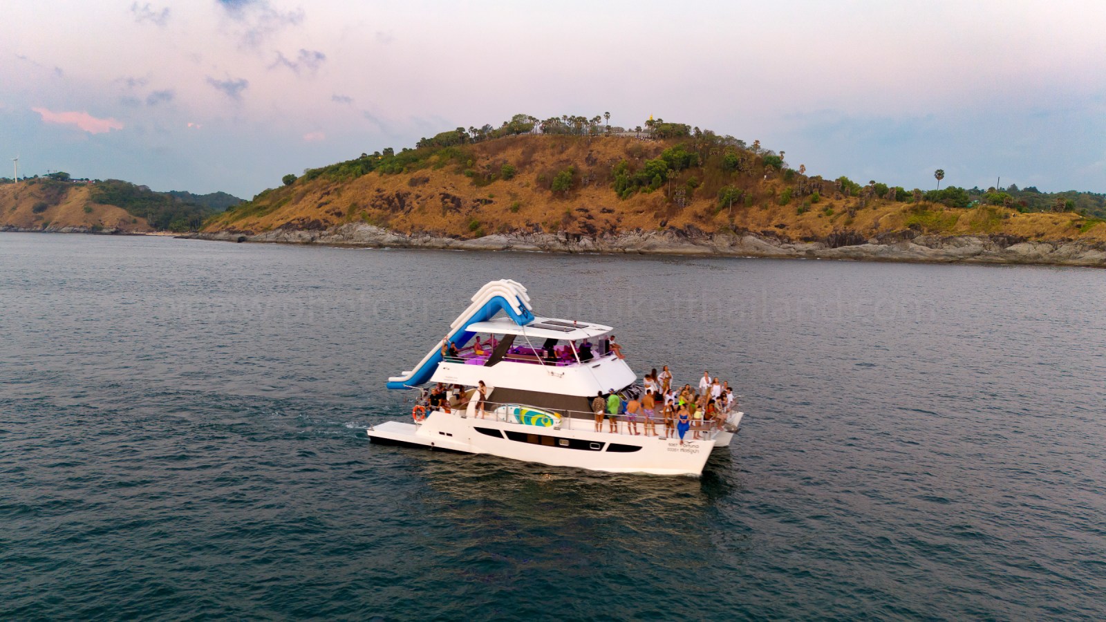 A group of people enjoying a party on a multi-level yacht with a water slide, anchored near a coastal area with hills in the background.