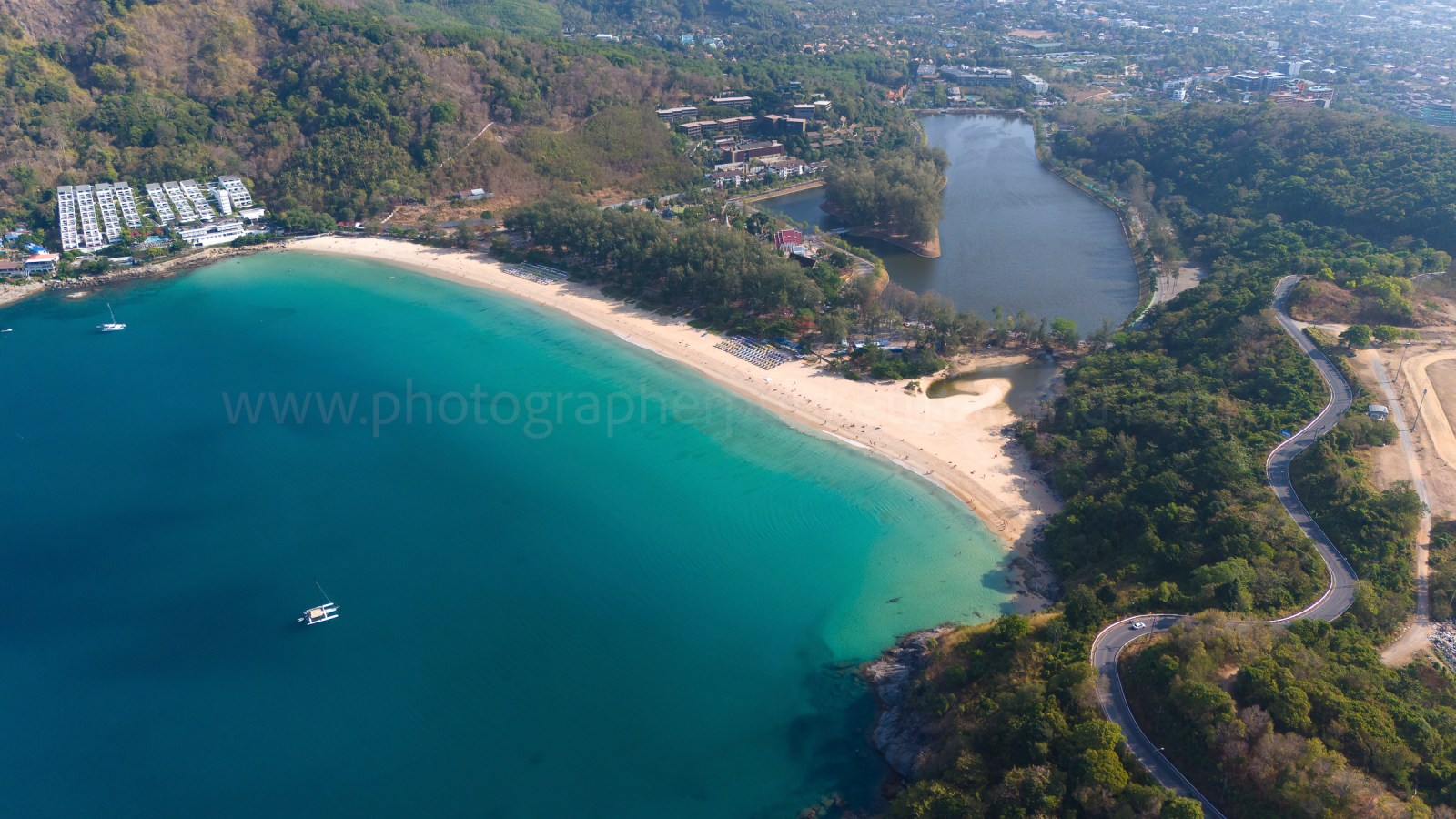 Aerial view of a serene beach with clear turquoise water, sandy shores, and greenery, featuring a sailboat and nearby resorts at naiharn beach phuket