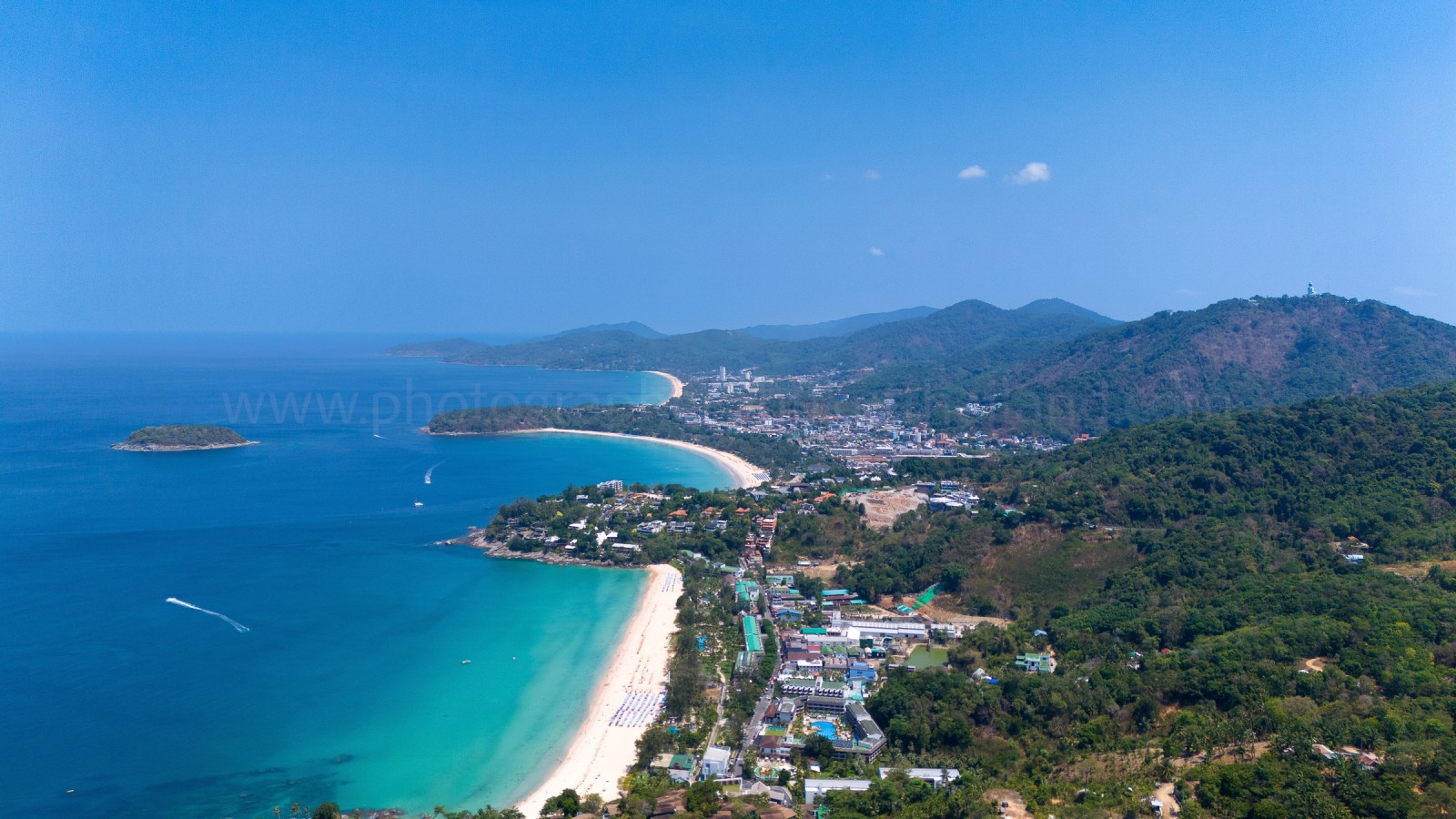 Aerial view of a coastal area featuring a sandy beach, turquoise waters, and a hillside town with greenery in Phang Nga Bay.