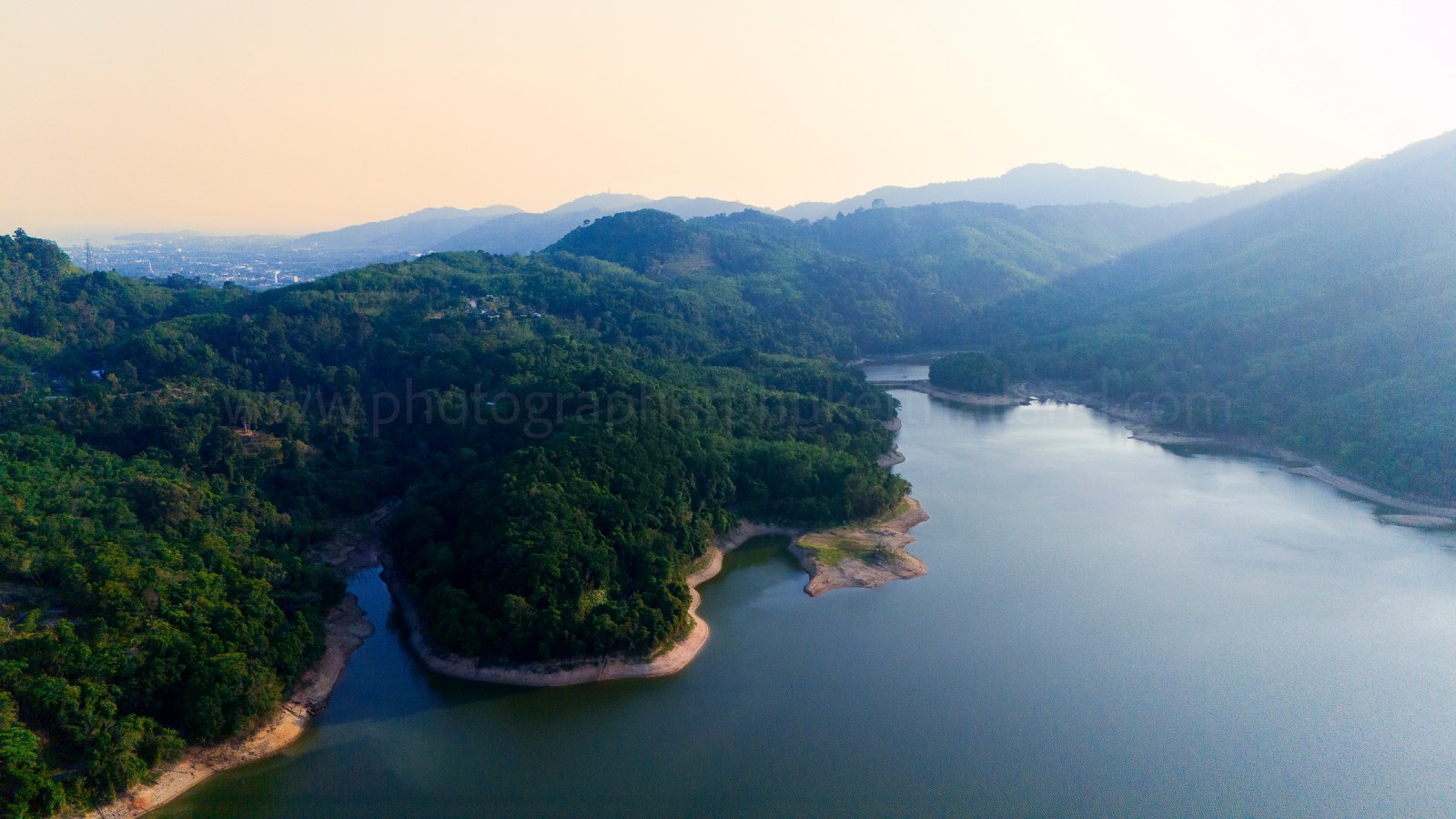 Aerial view of lush green hills surrounding a calm body of water, with a hazy horizon in the background.