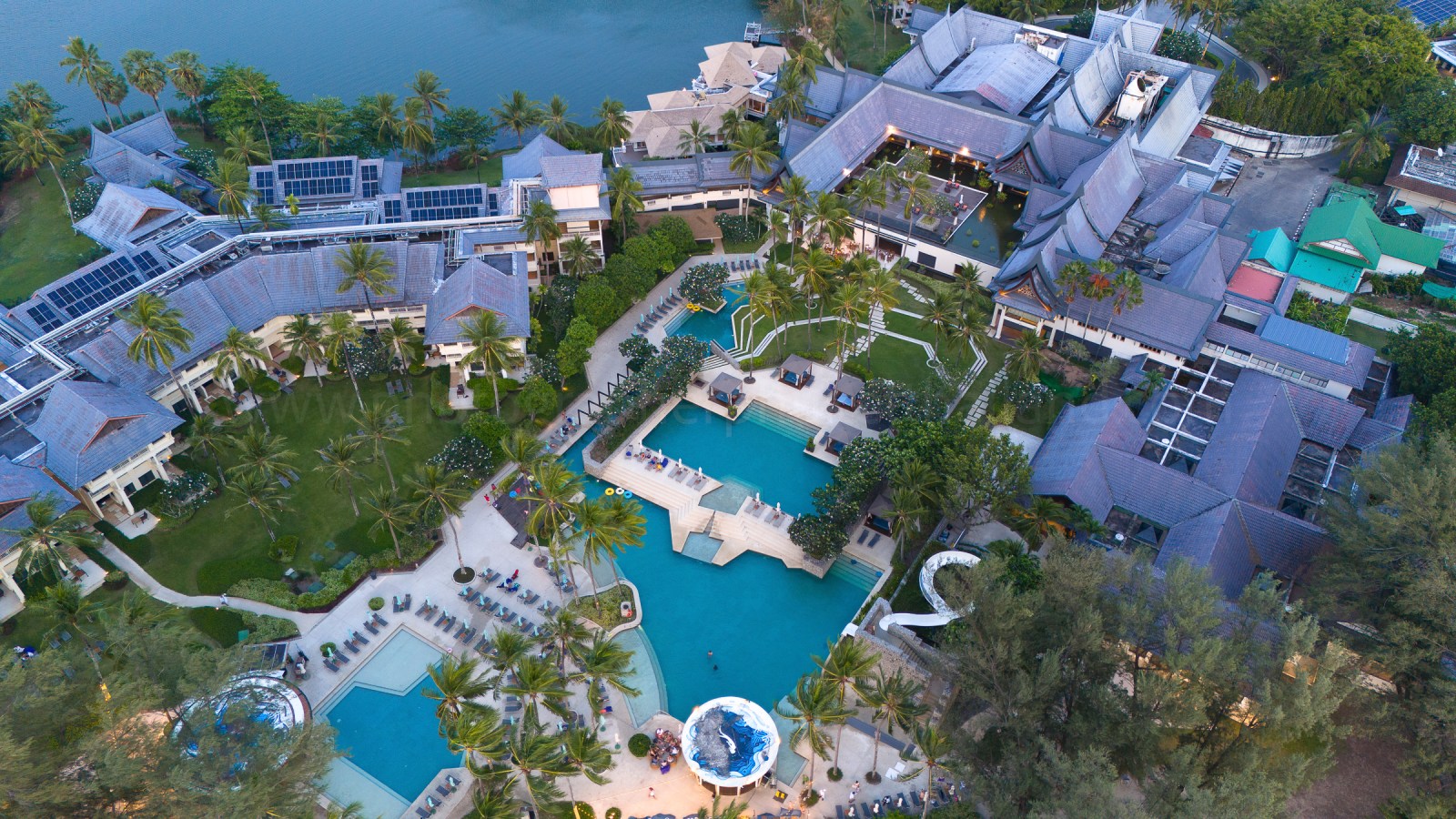 Aerial view of a resort complex featuring a large swimming pool, surrounded by palm trees and lush greenery.