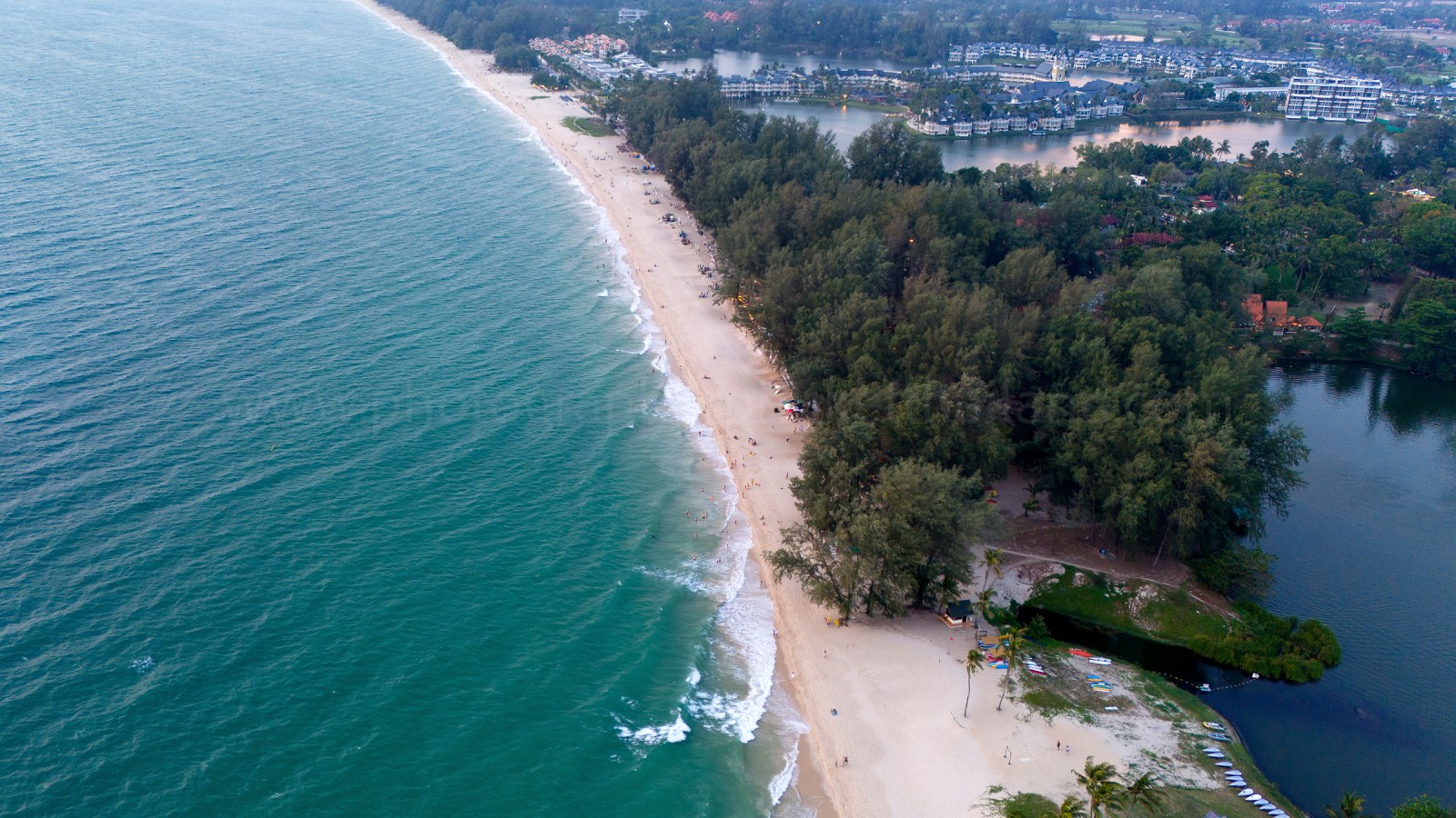 Aerial view of a sandy beach alongside a turquoise ocean, featuring a lush green area with trees and some beachgoers enjoying the sun.