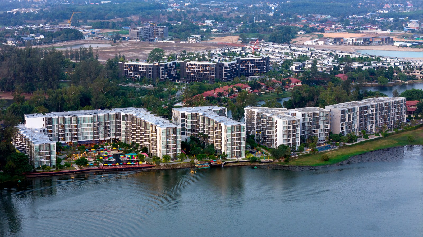 Aerial view of modern residential buildings alongside a calm water body surrounded by greenery.