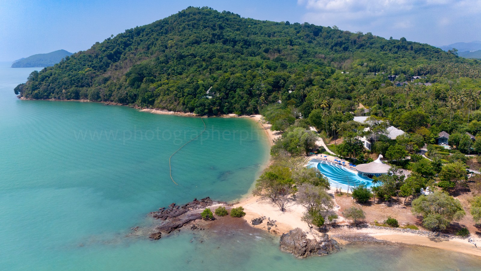 Aerial view of a serene beach with clear turquoise waters and lush green hills in the background, showcasing a swimming pool adjacent to the shore.