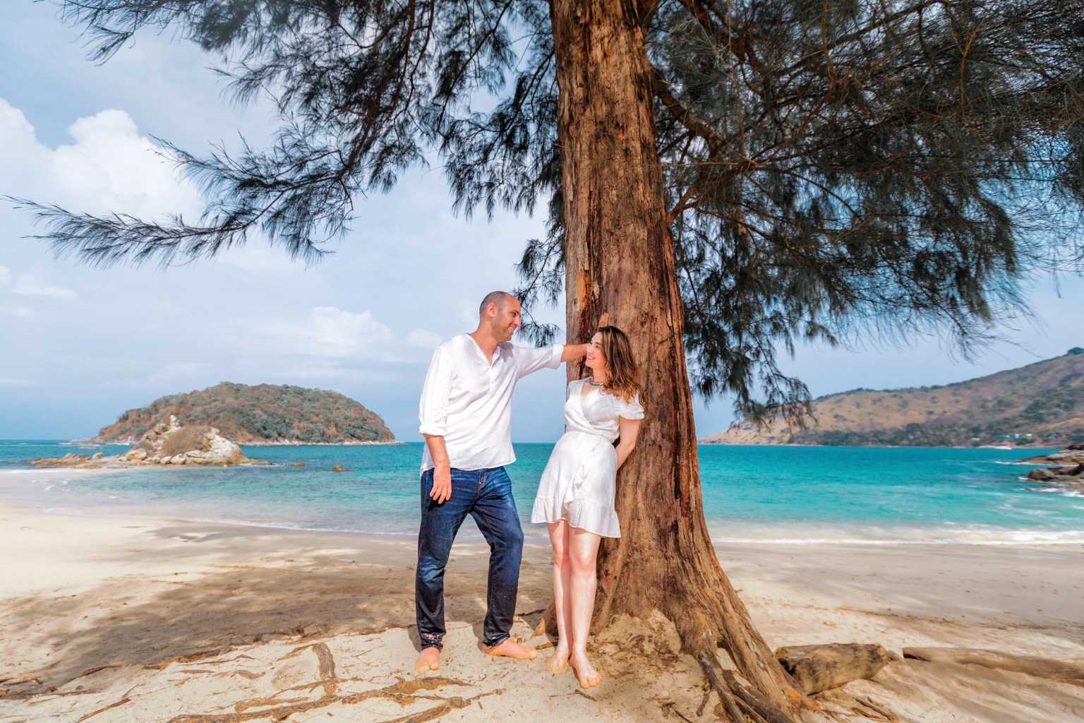 A couple posing happily together by a tree on a beach, with a beautiful turquoise sea and an island in the background.