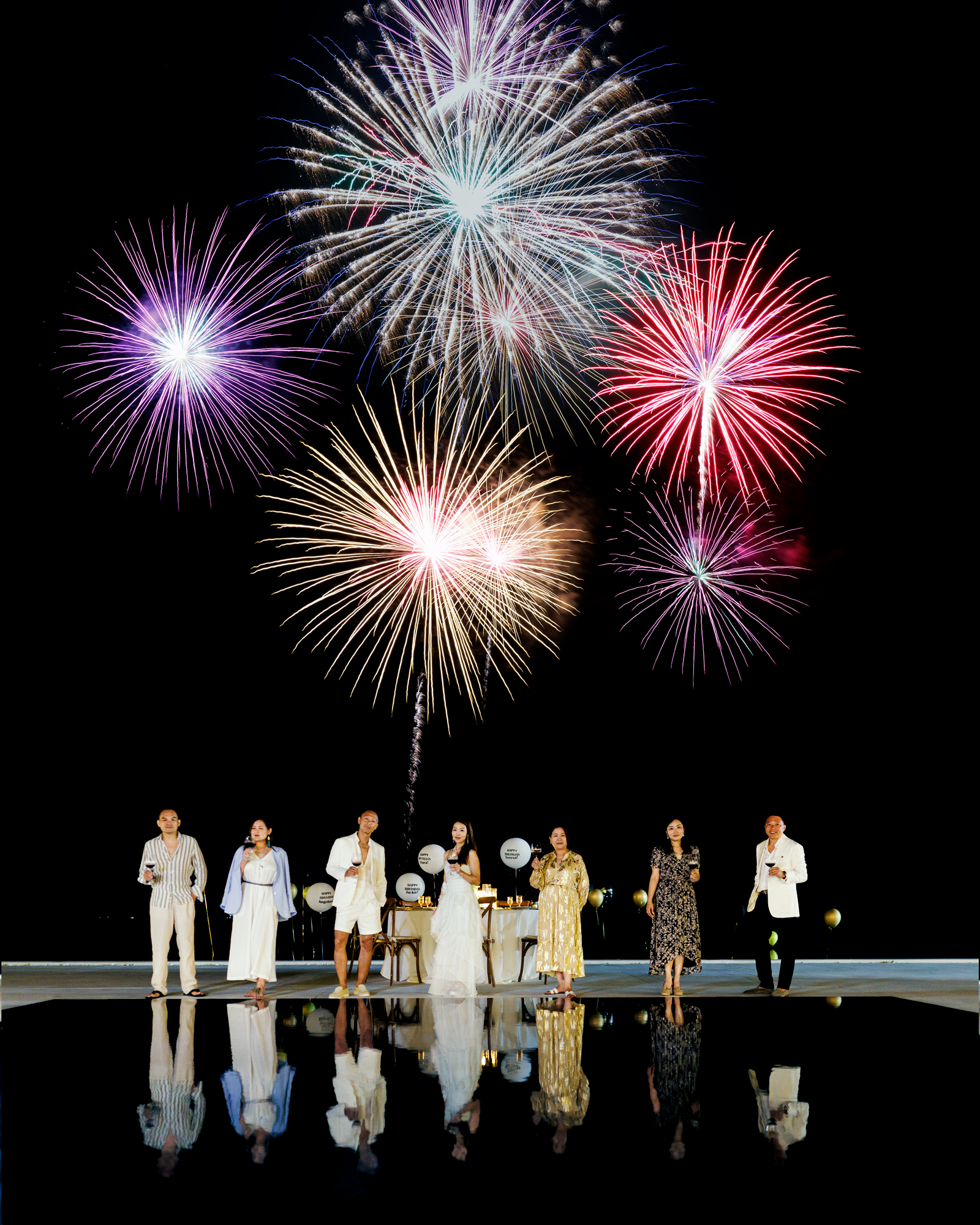 A group of five people standing in front of a table, celebrating with drinks as colorful fireworks explode in the night sky.