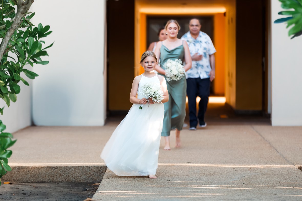 A young girl in a white dress holding a bouquet of flowers, walking in front of two women and a man along a pathway, with greenery on the sides.