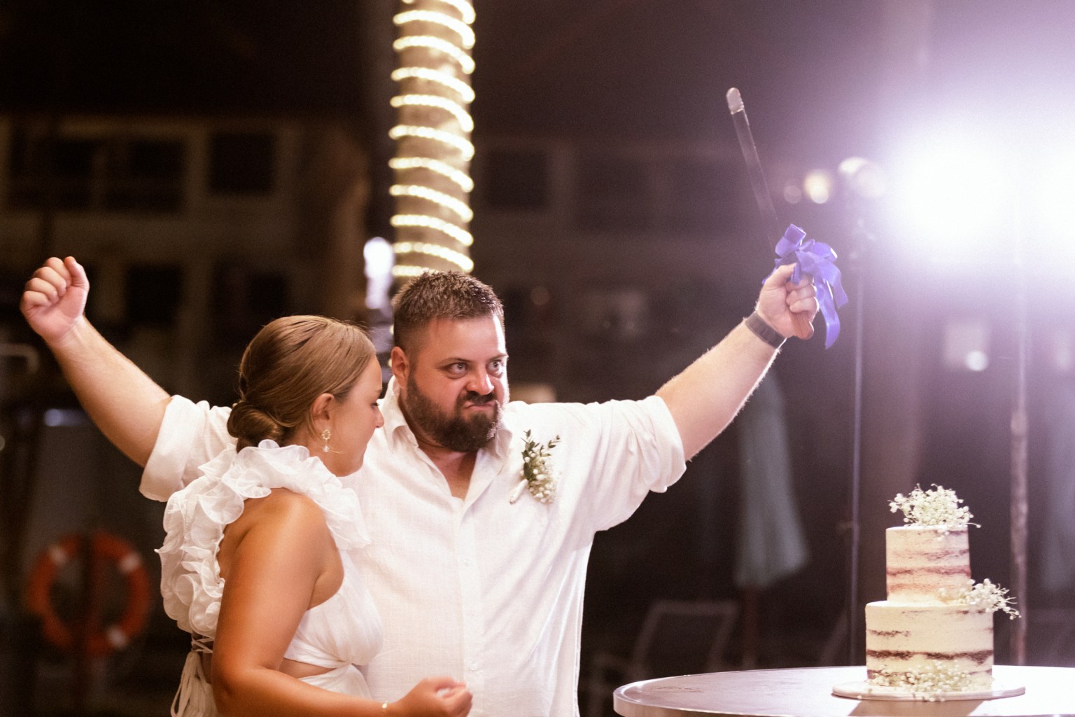 A bride and groom celebrate at their wedding reception, with the bride wearing a strapless white dress and the groom holding a ribbon cutting knife above a tiered wedding cake. The background features string lights, creating a festive atmosphere.