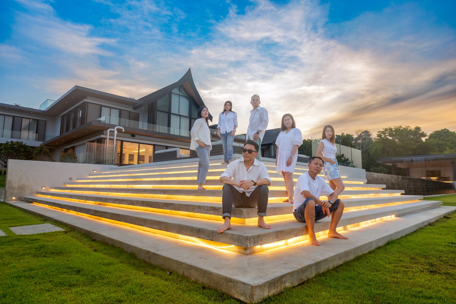 A group of people dressed in white outfits posing on illuminated steps outside a modern building during sunset.