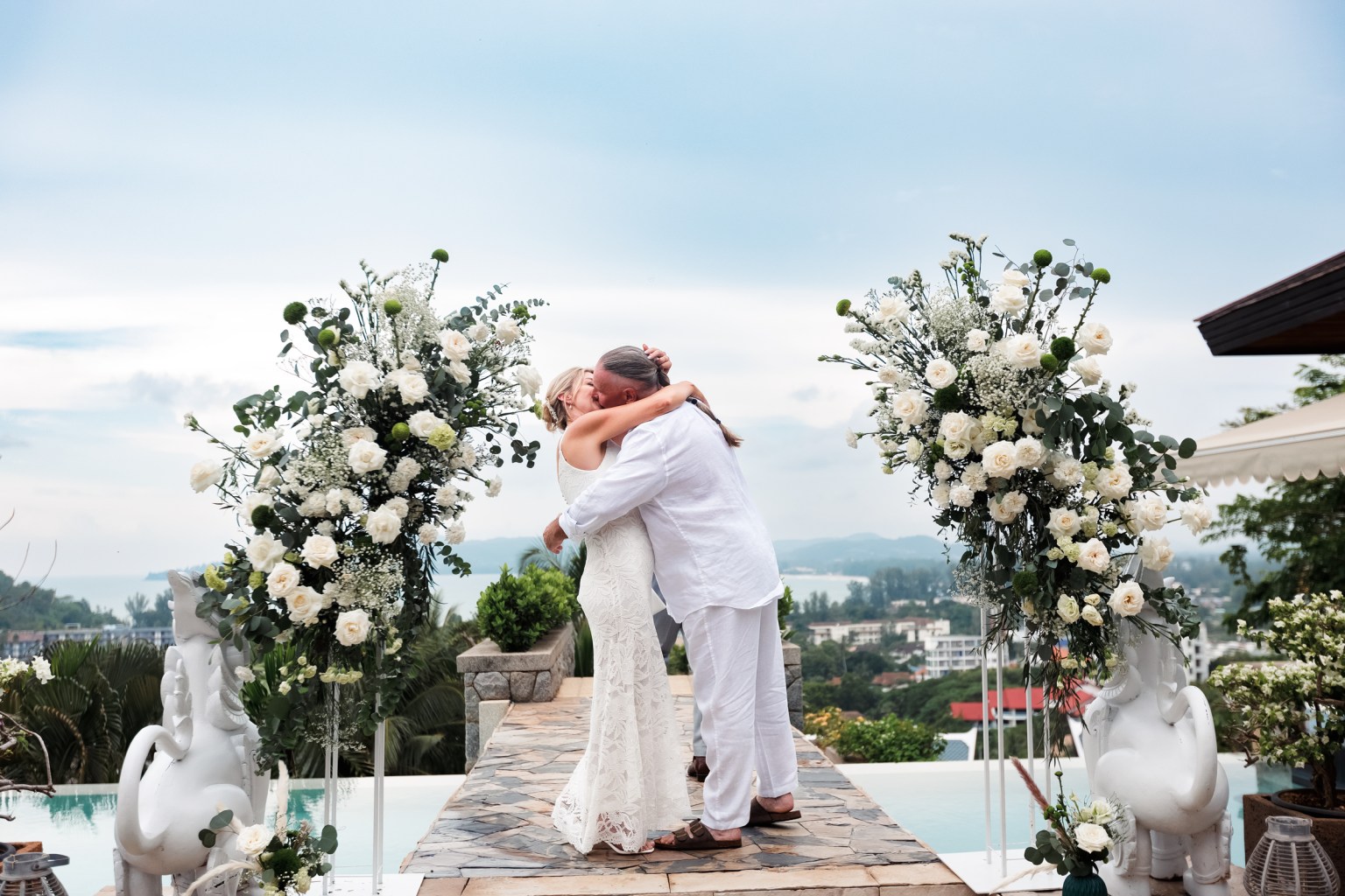 A bride and groom embrace during their wedding ceremony on a scenic outdoor setting, surrounded by floral arrangements and overlooking a beautiful landscape.