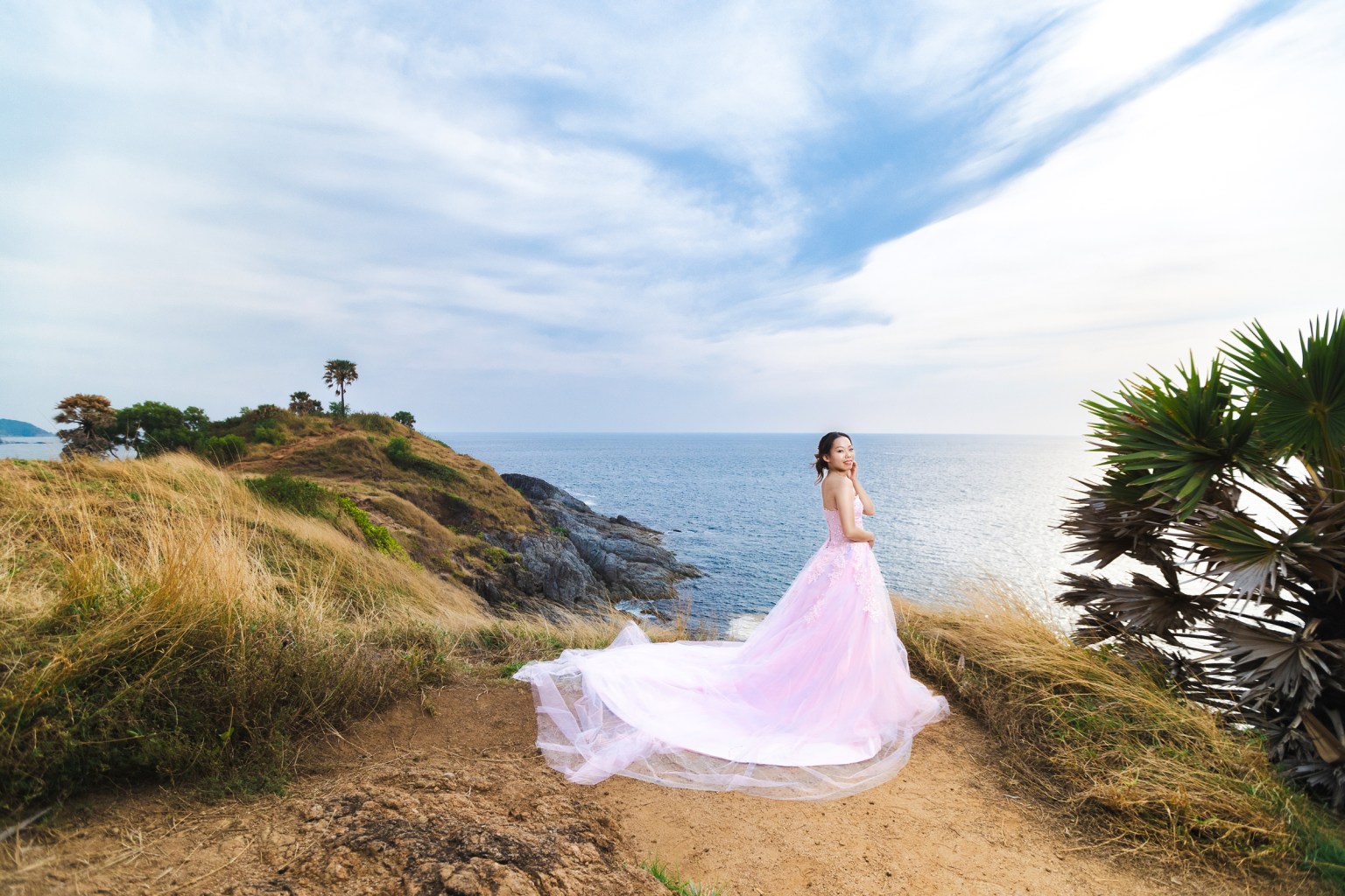 A woman in a flowing pink gown poses on a coastal cliff at promthep cape overlooking the ocean, with grassy terrain and palm trees in the background.