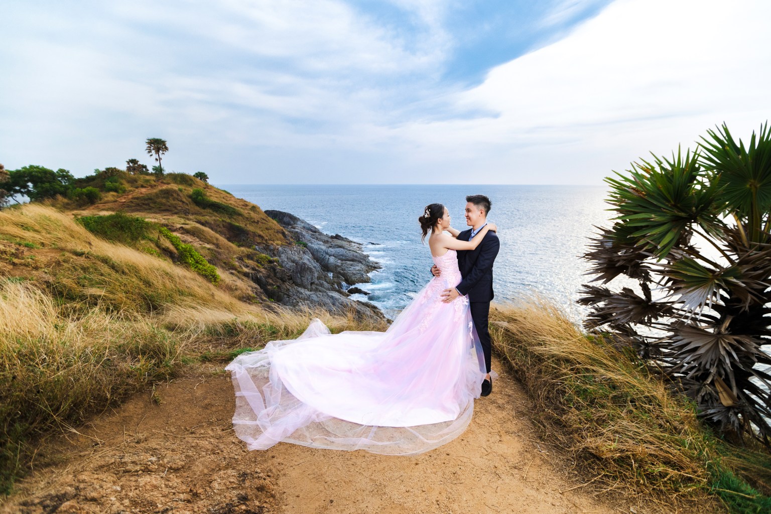 A couple embraces on a rocky cliff overlooking the ocean, with the woman in a long pink dress and the man in a dark suit.