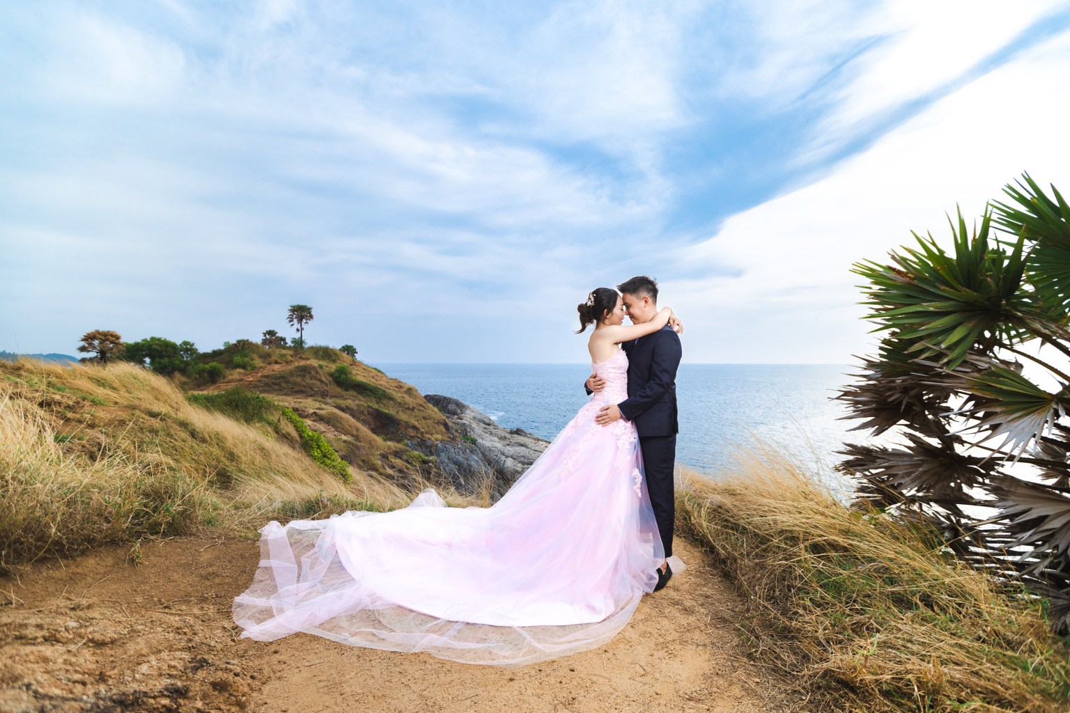 A couple embracing on the edge of a cliff at Promthep Cape, Phuket. The woman is wearing a flowing pink dress, while the man is in a black suit. The ocean and a partly cloudy sky serve as the backdrop.