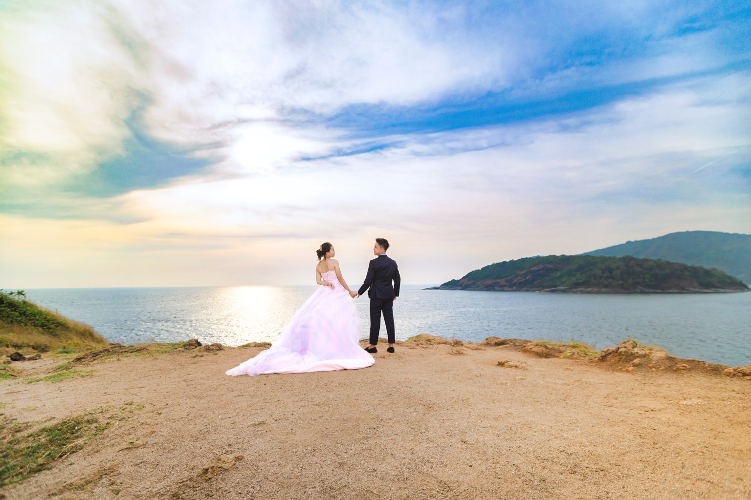 A couple holding hands while standing on a cliff overlooking the ocean at sunset, with a scenic mountainous island in the background.