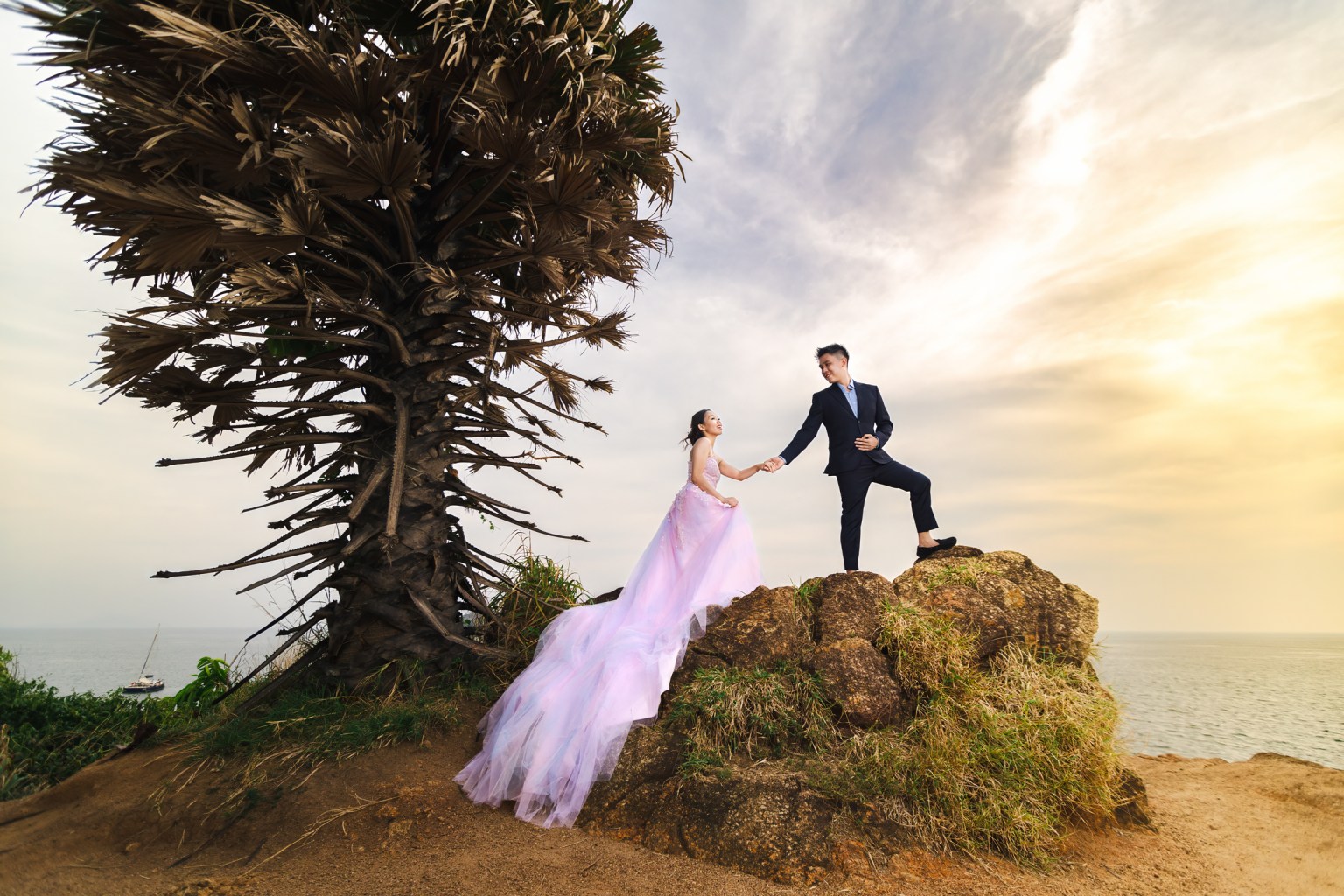 A couple standing on a rocky outcrop by the sea, with the man in a suit and the woman in a flowing pastel dress, holding hands against a sunset backdrop.