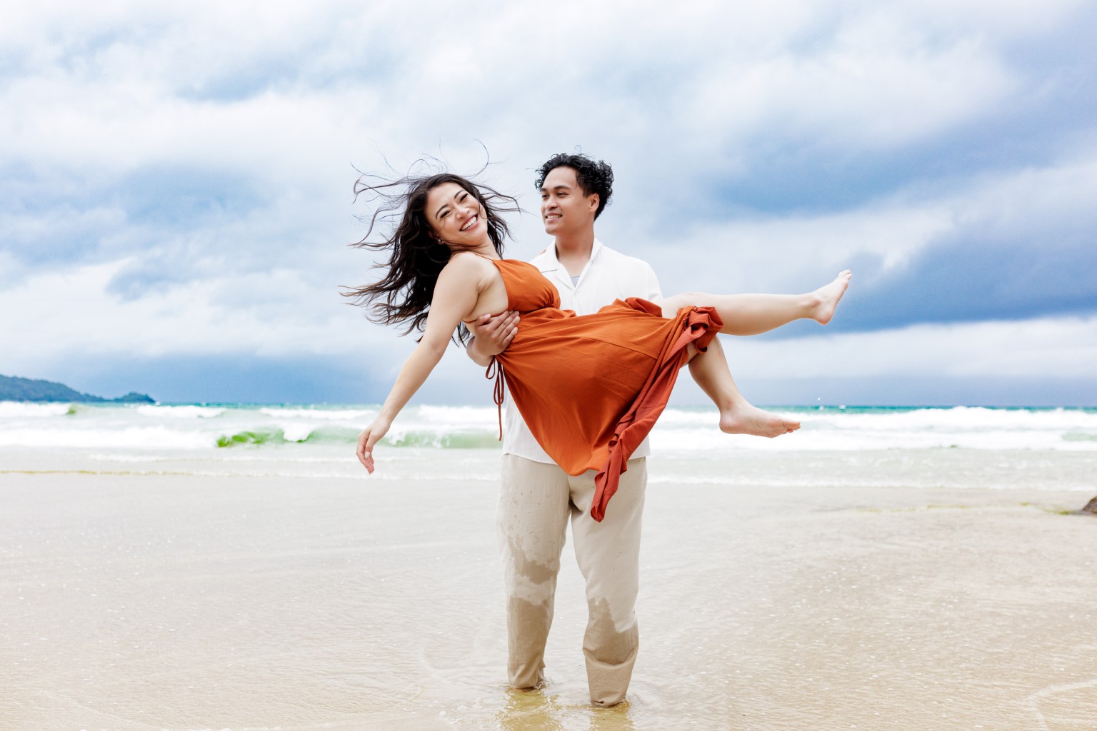 A couple joyfully posing on the beach, with the man lifting the woman dressed in an orange dress, against a backdrop of ocean waves and cloudy sky.