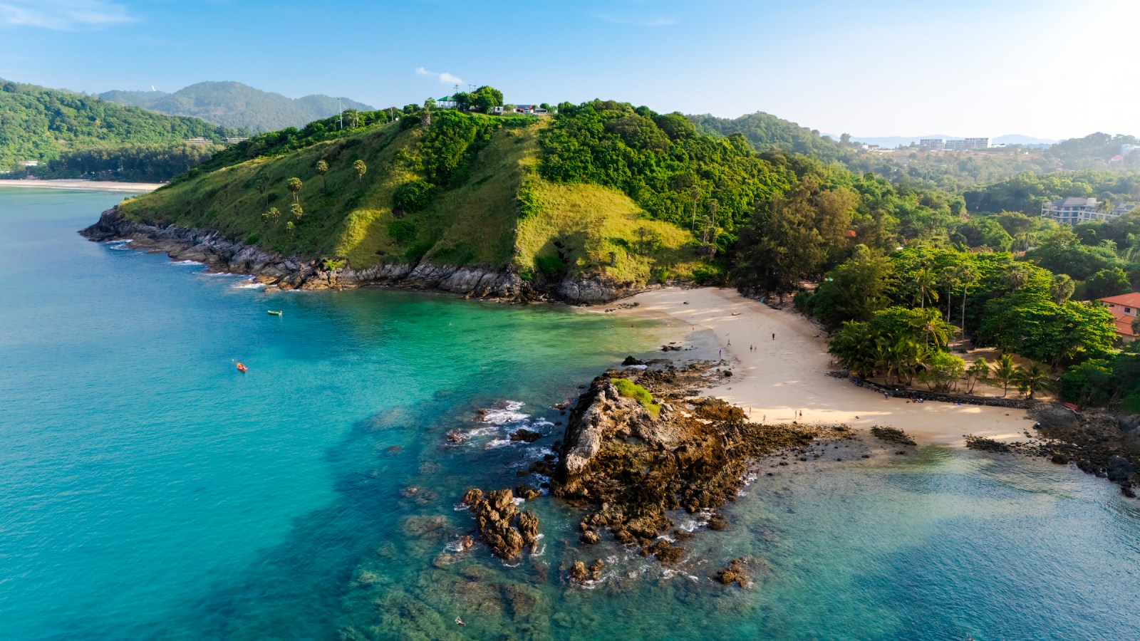 Aerial view of a lush green hill overlooking a clear blue sea with sandy beaches and rocky shores in the foreground.