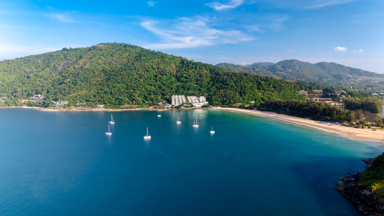 Aerial view of a serene beach with clear blue water and several sailboats anchored near the shore, surrounded by lush green hills and a few buildings overlooking the beach.