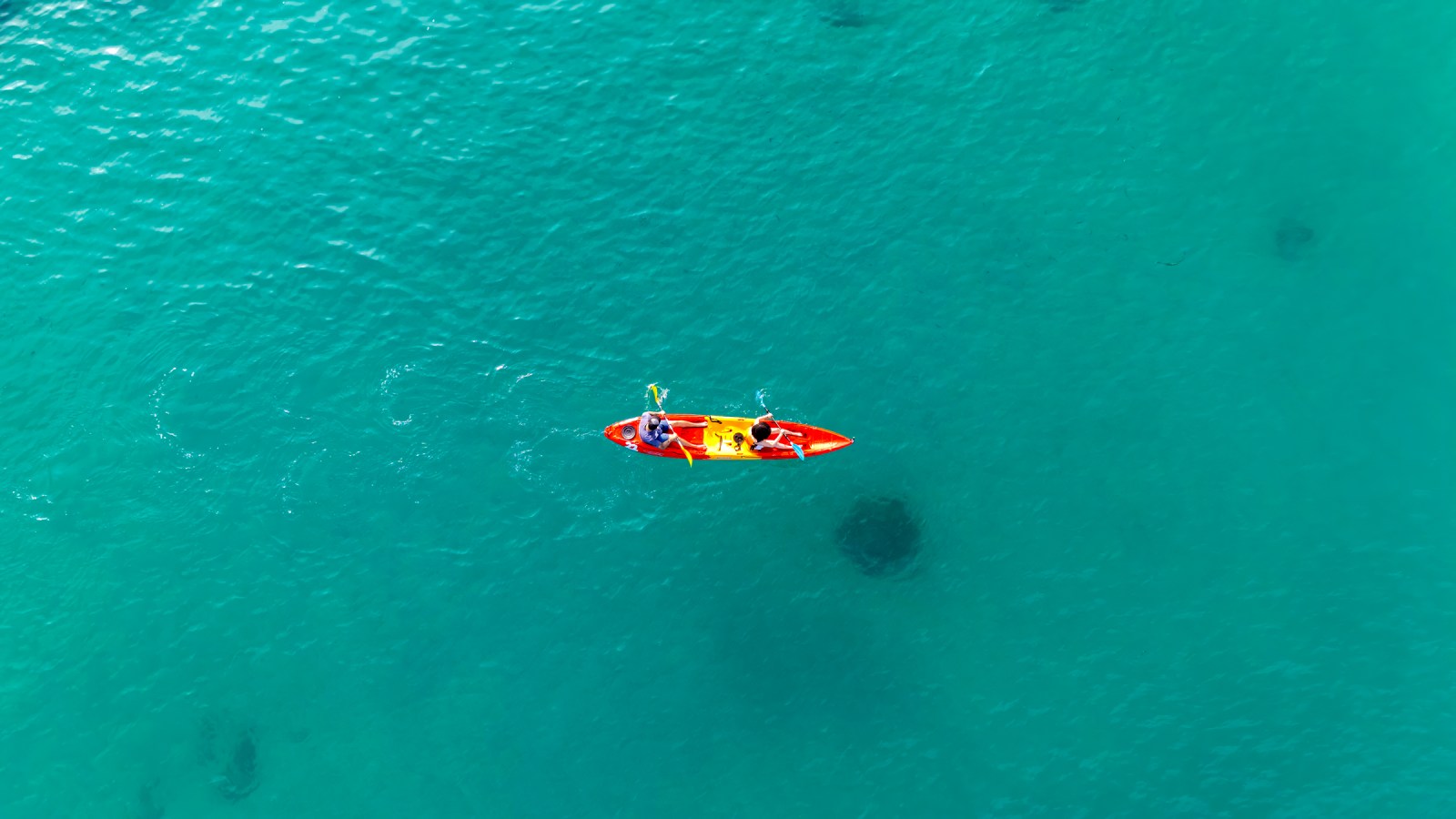 Aerial view of two people kayaking in a bright orange kayak on a turquoise sea.