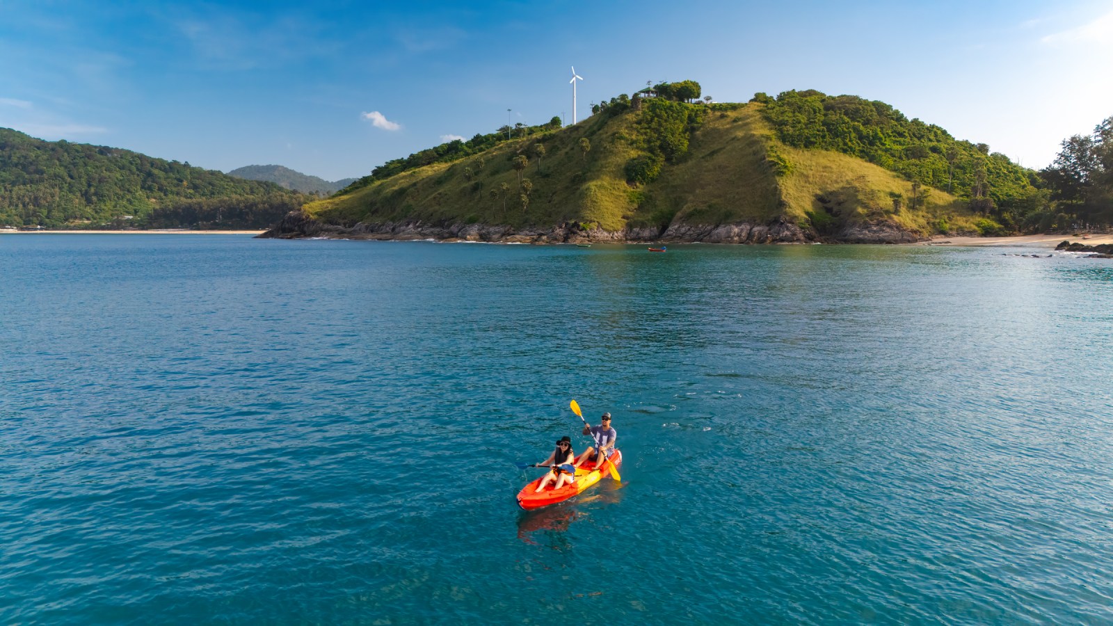 Two people kayaking on calm blue waters near a lush green hillside with a wind turbine in the background.