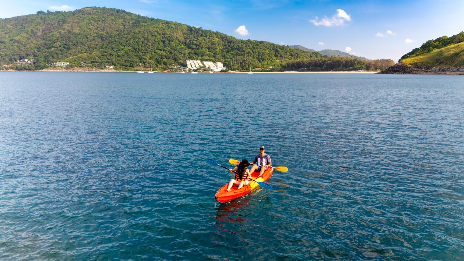 Two people kayaking on a serene blue lake surrounded by green hills and clear skies.