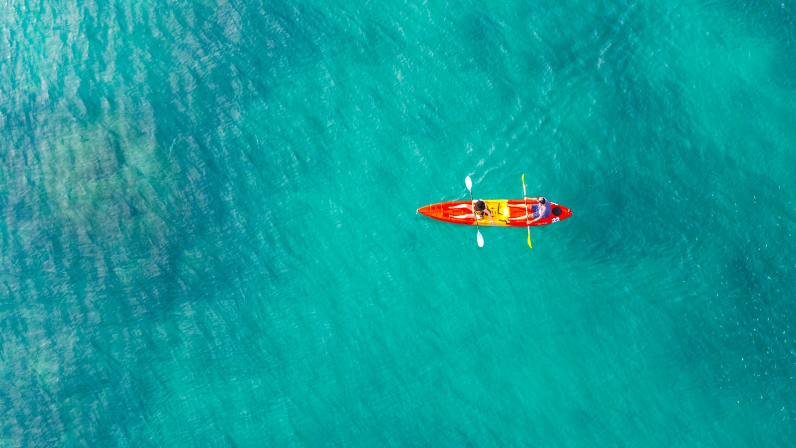 Aerial view of two people kayaking in a bright red kayak on clear turquoise water.