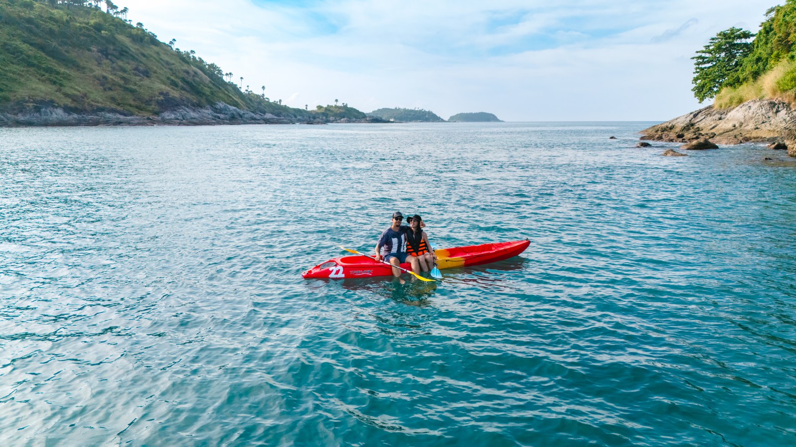 Two people sitting in a red kayak on a clear blue sea, with green hills and a cloudy sky in the background.