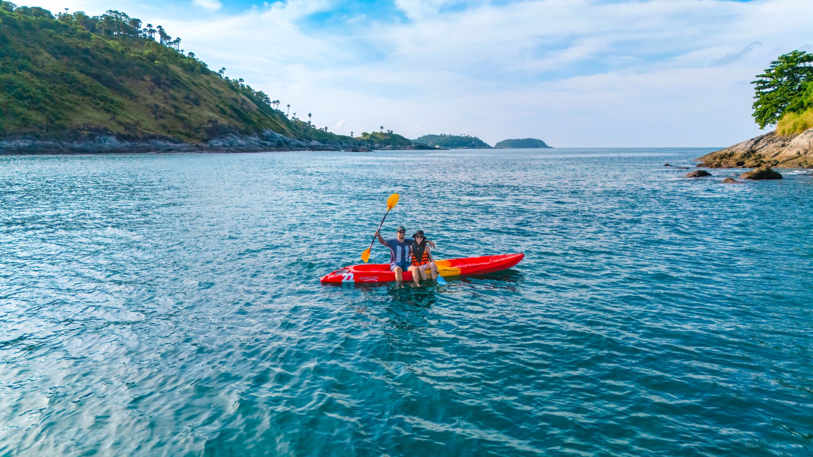 Two people in a kayak on clear blue water, surrounded by greenery and rocky shores.