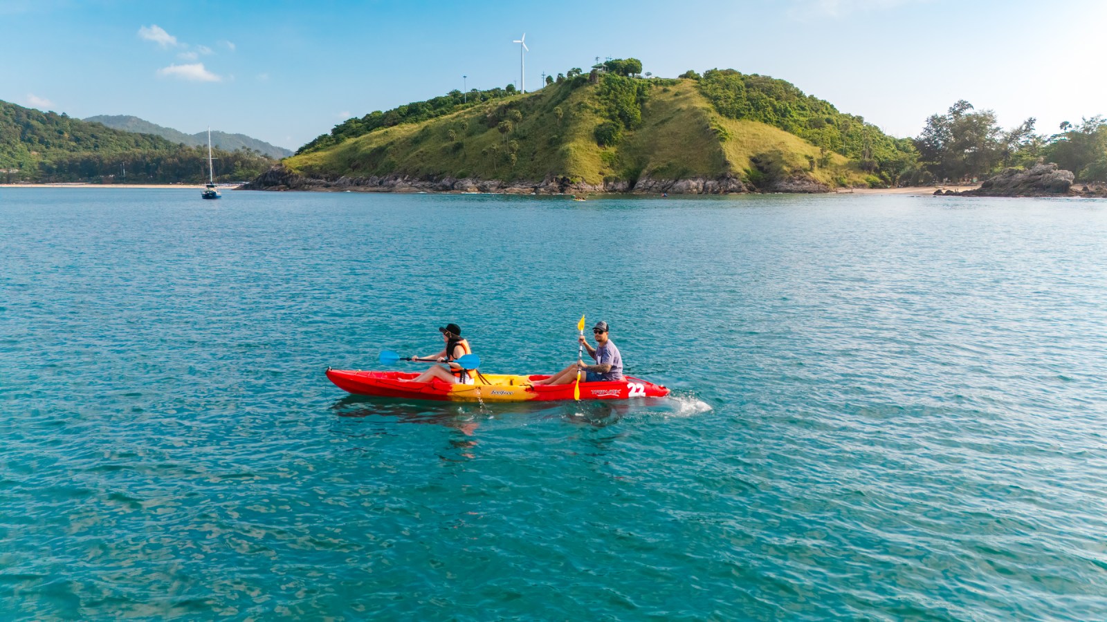 Two people kayaking in a bright red and yellow kayak on calm blue waters, with green hills in the background and a sailboat in the distance.