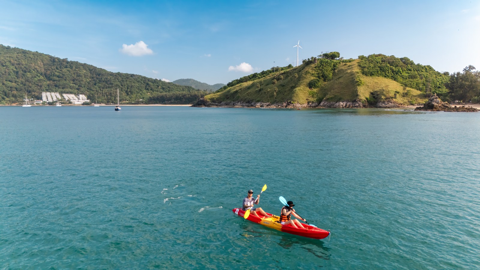 Two people kayaking in a calm body of water with a green hillside in the background under a blue sky.
