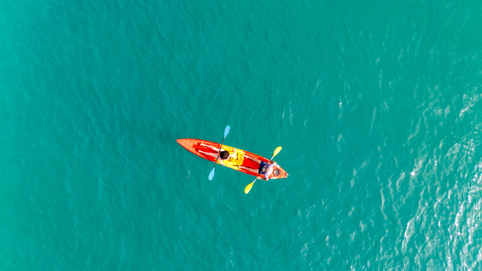 Aerial view of two people kayaking in a bright orange and yellow kayak on clear turquoise water.