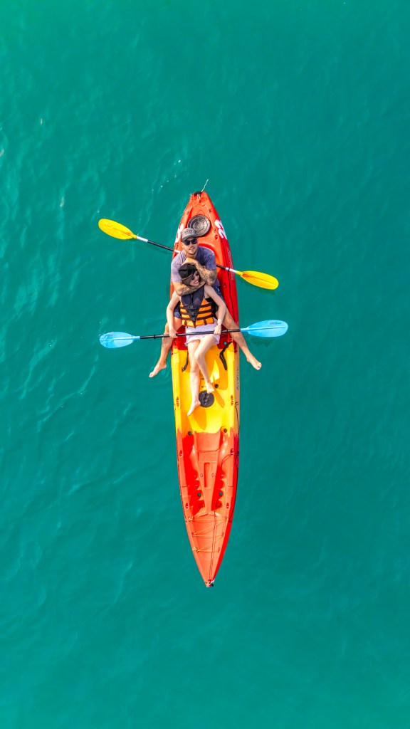 Aerial view of three individuals kayaking in a vibrant orange and red kayak on clear turquoise waters.