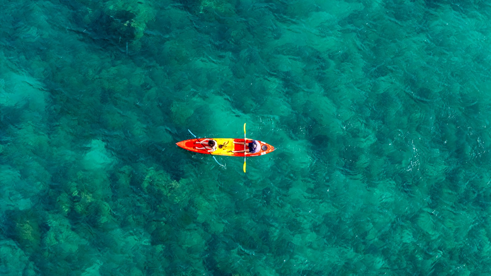 Aerial view of two people in a bright red kayak with yellow accents, paddling through clear turquoise waters.
