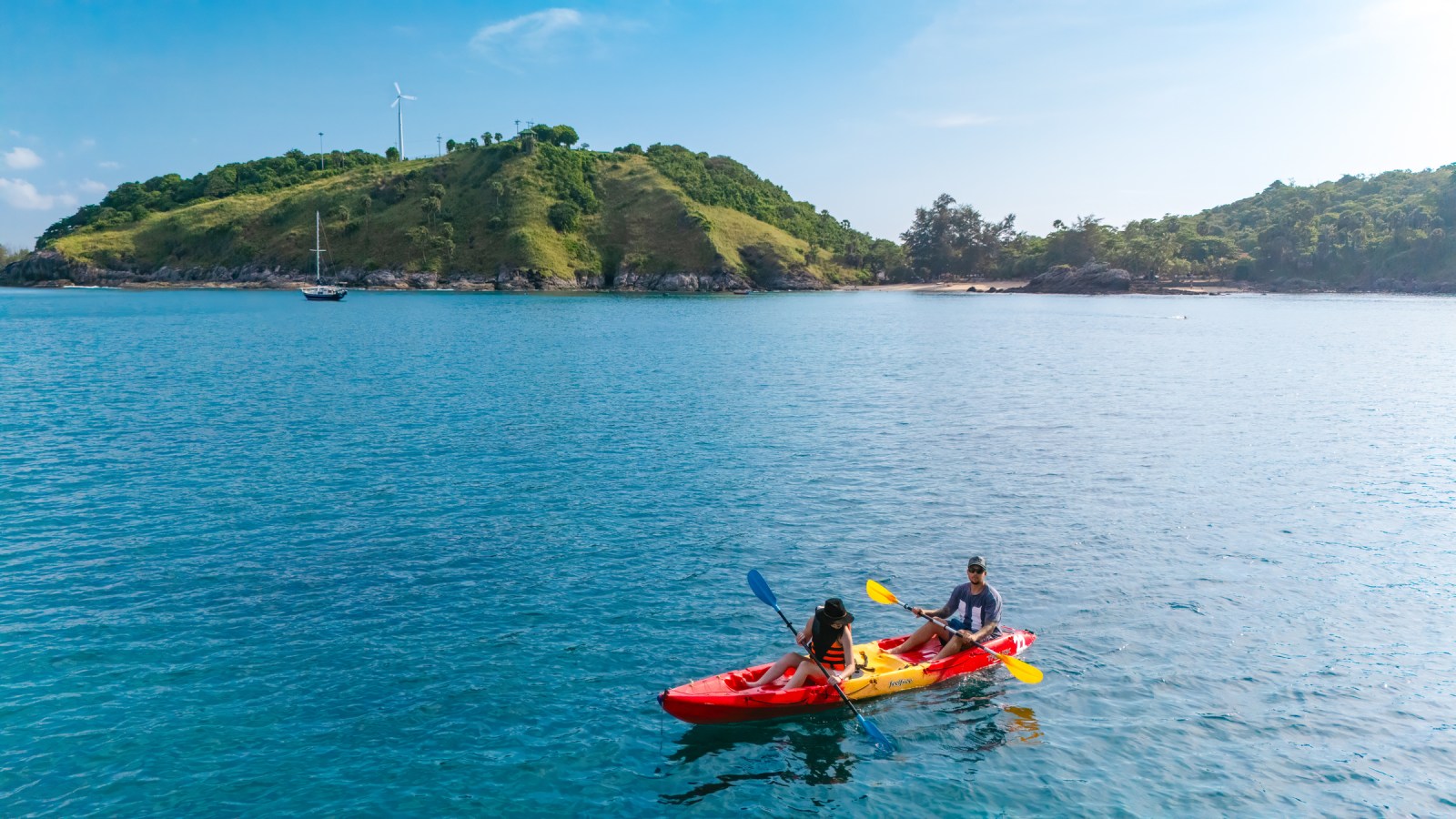 Two people kayaking on calm waters with a green island and wind turbines in the background.