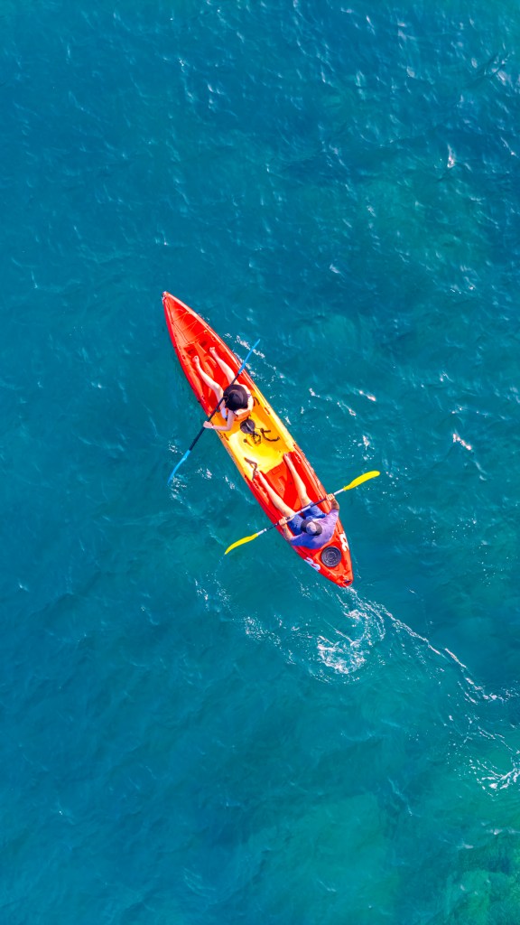 Aerial view of two people kayaking in a bright orange and yellow kayak on clear blue water.