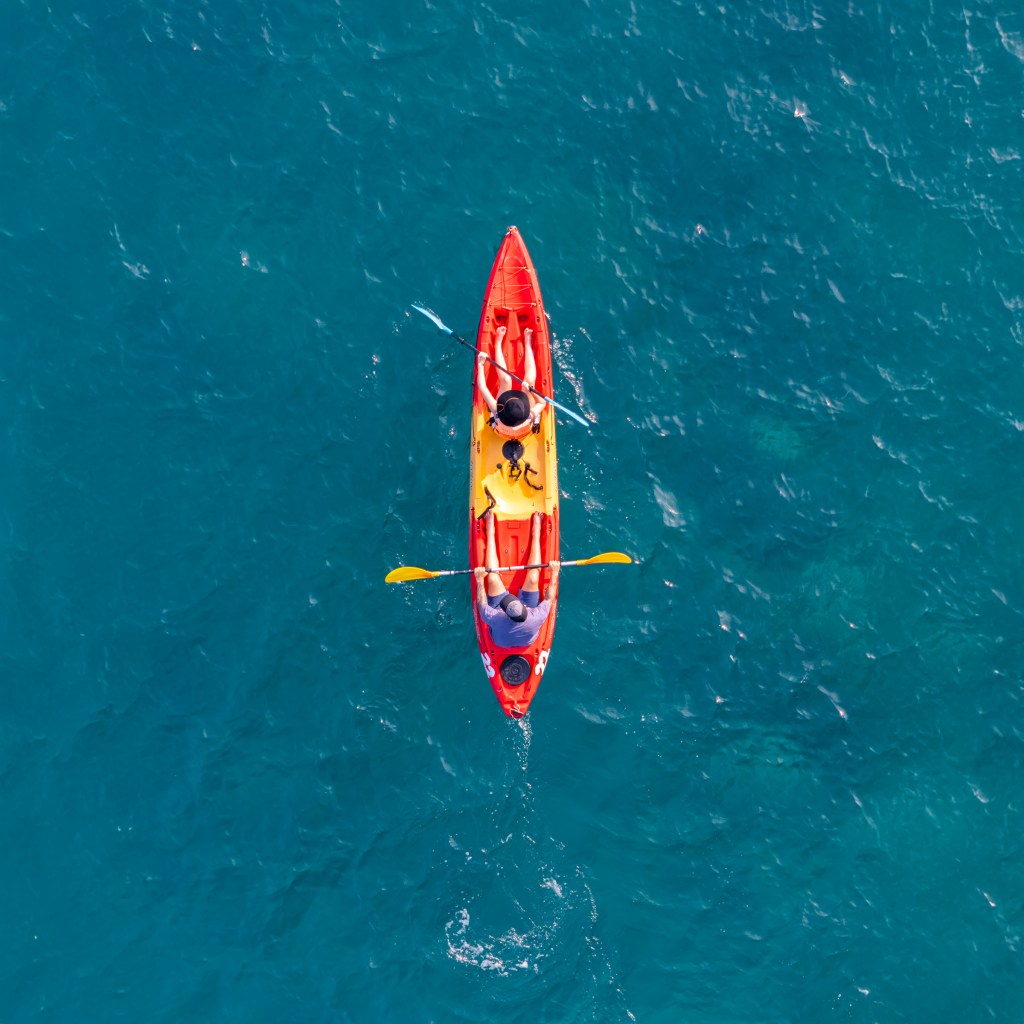 Aerial view of two people paddling in a bright red and yellow kayak on clear blue water.